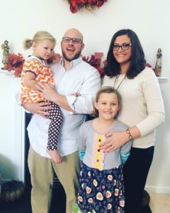 "Dr. Haley Perry, owner of Elite Eye Care, with her family. On the left, her husband Aaron holds their youngest daughter, while their oldest daughter stands in front of Dr. Perry. The joyful family poses in front of a fireplace adorned with fall leaves, beaming with smiles."