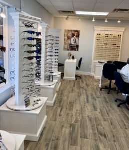 Elite Eye Care Arden NC optical image showcases glasses towers filled with a wide selection of eyeglasses and sunglasses. The optical area features a modern farmhouse style decor, faux wooden flooring, and blue-gray walls, creating a warm and inviting atmosphere for visitors.