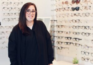 Marissa Gehring, technician at Elite Eye Care in Arden, NC, stands at the rock-covered front desk adorned with a Monstera plant. The background features a blue wall, creating a welcoming atmosphere.