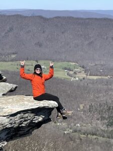 Dr. Casey Krug, Associate Optometrist at Elite Eye Care in Arden, NC, enjoying a hike. In the image, she is sitting on the ledge of an overlook, surrounded by stunning mountains and valleys. Dr. Krug is wearing a black beanie, stylish sunglasses, an orange coat, and comfortable black pants paired with hiking boots. Her adventurous spirit shines through as she takes in the breathtaking scenery
