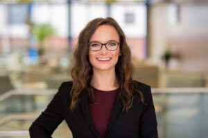 Dr. Casey Krug, Associate Optometrist at Elite Eye Care in Arden, NC. She is wearing a black blazer and black glasses, with wavy brown hair. Dr. Krug is standing, smiling confidently in front of a blurred background