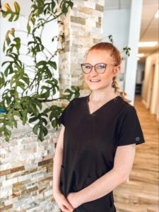 Kirsten McCarthy, technician at Elite Eye Care in Arden, NC, stands confidently at the rock-covered front desk. A vibrant Monstera plant gracefully grows up the desk, adding a touch of greenery to the scene. The background features a soothing blue wall, creating a pleasant and welcoming atmosphere.