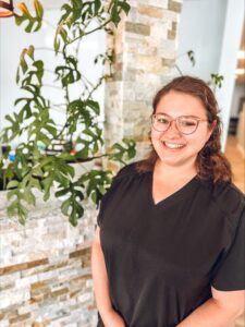 Raven Bowes, Office Administrator at Elite Eye Care in Arden, NC, stands confidently at the rock-covered front desk adorned with a beautiful Monstera plant climbing up its side. The vibrant blue wall serves as a stylish background, complementing the professional and welcoming atmosphere.