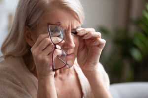 Photo of lady with closed eyes. She is holding her eyes with her fingertips and is holding a red pair of glasses. She looks tired/frustrated.