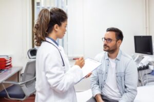 Female doctor in white coat talks to male patient before procedure. Doctor is holding a clipboard and pen. 