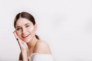 Brunette female in front of white background putting sunscreen on her face and smiling